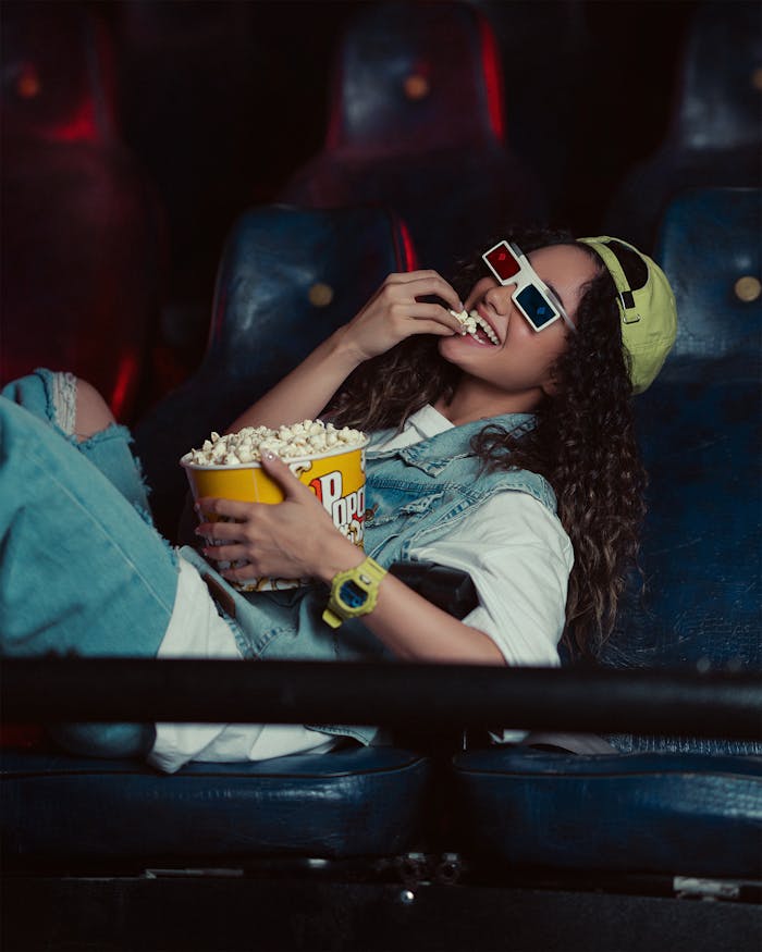 about-02 Woman with long hair and colorful attire enjoying popcorn in a cinema wearing 3D glasses.